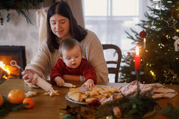 Happy mother with cute baby son making gingerbread christmas cookies together, decorating with sugar glaze against christmas tree. Christmas family traditions, winter holiday preparation