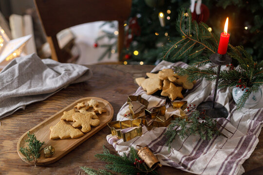 Making holiday cookies. Christmas gingerbread cookies on plate, fir branches, candle and metal cutters on rustic table against decorated christmas tree in evening. Holiday preparation