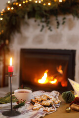 Merry Christmas! Gingerbread christmas cookies, warm cup of tea, fir branches and ornaments on table on background of festive decorated fireplace with lights in eve. Cozy winter time