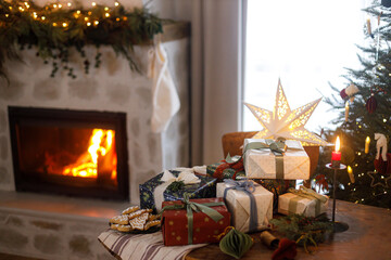 Merry Christmas! Stylish wrapped christmas presents, ornaments, candle, gingerbread cookies on table on background of festive decorated fireplace and christmas tree with lights. Holiday eve