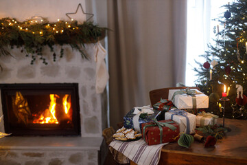 Stylish christmas presents, ornaments, gingerbread cookies on table on background of decorated fireplace and christmas tree lights in nordic living room in magical eve. Merry Christmas!