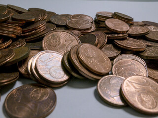 A close-up photograph of a scattered pile of copper-colored Euro cent coins, predominantly 1 and 2 cent denominations, lying on a light, plain surface. 