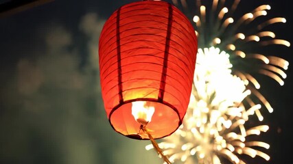 A vibrant red paper sky lantern glowing warmly as it floats into the night sky with a spectacular fireworks display celebrating a festive occasion - Powered by Adobe