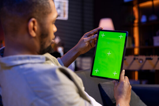 Black freelancer uses green screen mockup on modern tablet to work in the living room, sitting at home with the gadget and solving tasks. Relaxed casual young adult in his trendy space.