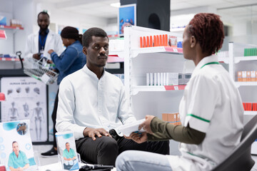 Black healthcare assistant offers personalized medication advice to a young customer in a modern stacked pharmacy shop, recommending vitamins and drugs for effective self care.