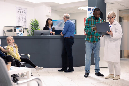 Elderly female physician in lab coat consults with male patient in hospital waiting room. Senior doctor with laptop discusses recovery plan with black man as receptionist assists clinic visitors.