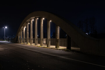 Fototapeta premium Night view of a concrete arch bridge illuminated by streetlights, with strong shadows and an empty road creating a calm urban atmosphere.