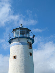 Close-up of a coastal lighthouse painted white and blue, illuminated by sunlight against a bright blue sky with scattered clouds. The image conveys calmness, guidance, and maritime landscapes.