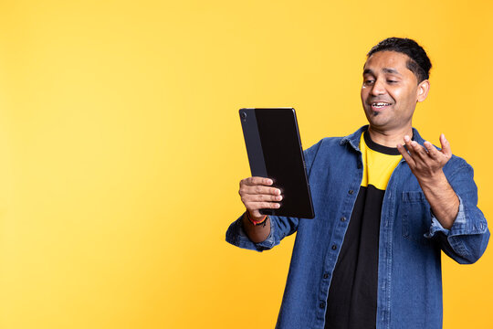 Indian person using tablet to call his friends on remote video conference, waving hand and standing against studio background. Young guy laughing during an online web meeting call. - Powered by Adobe