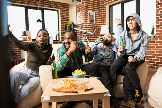 Cheerful group of friends sitting in living room, sharing pizza, snacks and drinks. Diverse men and women gather in apartment, watching game on TV and reacting with excitement after team victory. - Powered by Adobe