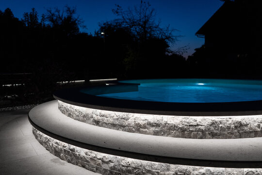A modern outdoor pool glowing at night with vibrant LED lighting inside and around the water, creating a luxurious, serene, and atmospheric scene ideal for relaxation and nightlife visuals.