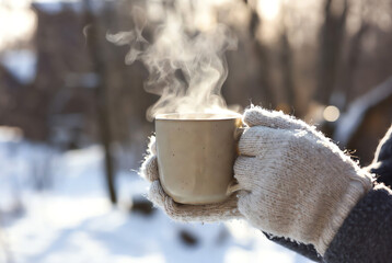 Person in warm knitted gloves holding a steaming mug outdoors in a sunny snowy winter landscape, offering comfort and cozy warmth.