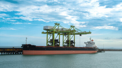 Fototapeta premium Large cargo ship docked at a pier beneath massive green port cranes set against a blue cloudy sky. Transportation and logistics