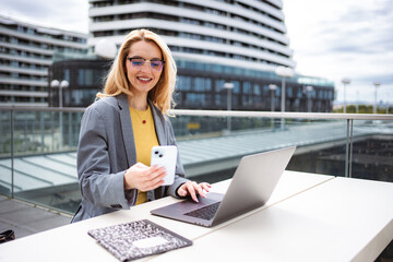 Businesswoman working remotely outdoors, using smartphone and laptop