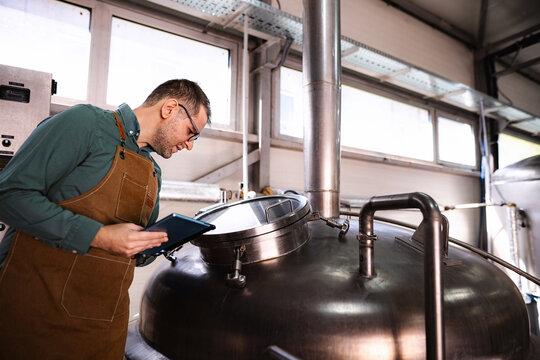 Man wearing apron checking beer fermentation on tablet in microbrewery