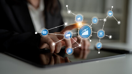 A businesswoman uses a touchscreen device to interact with digital icons representing growth and analytics in a modern office setting, highlighting technology impact on business. Tessel