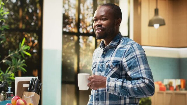 Portrait of joyous african american man drinking his morning coffee in a mug before eating bread and croissants for breakfast. Calm morning surrounded by kitchenware and healthy food. Camera B.
