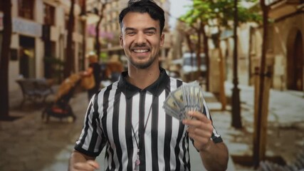 Young hispanic man in referee uniform holding us dollars and smiling triumphantly in a lively outdoor city street showcasing excitement and success. - Powered by Adobe