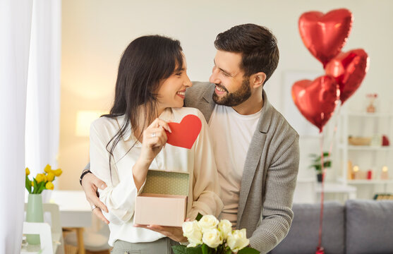 Happy young couple celebrating birthday or Valentine's Day at home. Smiling loving husband makes romantic holiday surprise for wife giving her present, red heart shaped card and bouquet of white roses