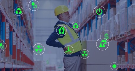 Leaning worker holding back in long pallet aisle, wearing yellow hard hat and vest with icons