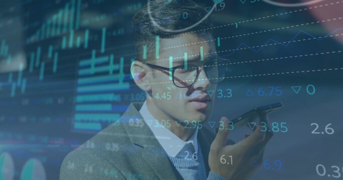 Talking Asian businessman using smartphone at desk viewing overlaid charts in suit and eyeglasses
