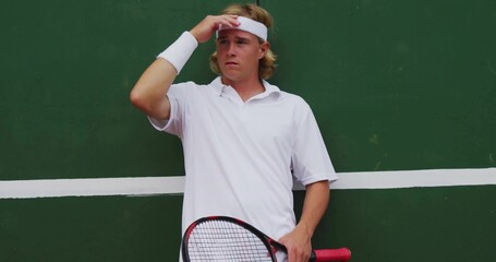 Shielding male player squinting at green practice wall, holding red racket, white bands, white polo