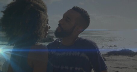 Naklejka premium Embracing couple leaning at rocky shoreline with ocean horizon, sun flare, tank top and tie-dye tee