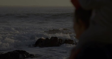 Watching woman wearing jacket, hat gazing at breaking waves on rocky shore at dusk, copy space