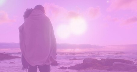 Standing couple wrapped in light blanket facing ocean on rocky shore, pink-purple sun, copy space