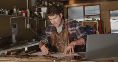 Leaning mature craftsman wearing leather apron marking plans at wooden workbench with open laptop