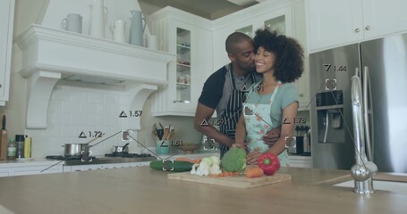Embracing couple wearing aprons preparing meal at kitchen island, wooden cutting board and veggies