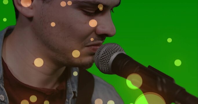 Leaning singer singing into mic stand in studio with green backdrop, denim jacket over maroon shirt