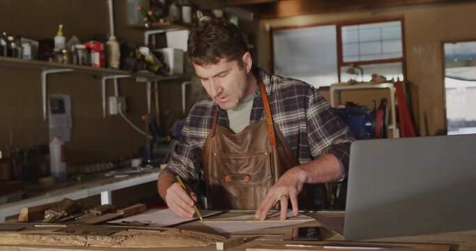 Leaning mature craftsman wearing leather apron marking plans at wooden workbench with open laptop