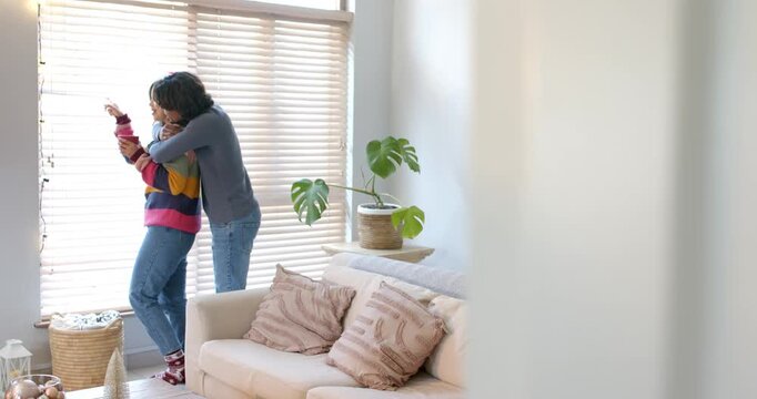 Diverse couple at home embracing sipping red mug as partner entering guiding blinds kissing neck
