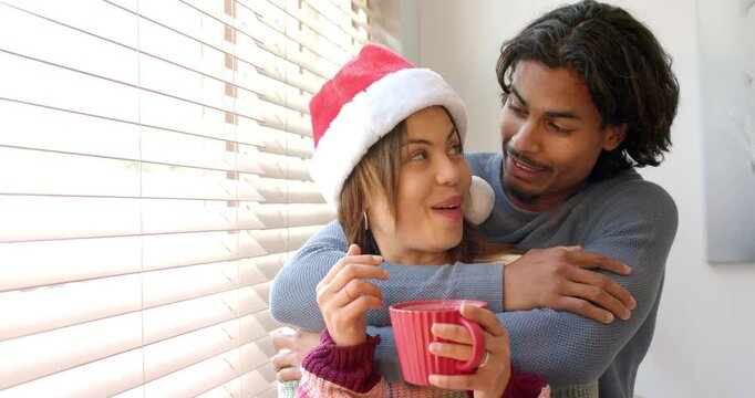 Curious diverse couple in Santa hat parting blinds at window, peeking, cuddling and sipping red mug