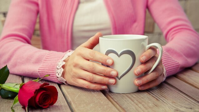 Woman holding a heart mug, cozy moment with a red rose on a wooden table, symbolizing love, romance, and a warm beverage for valentine's day morning - Powered by Adobe
