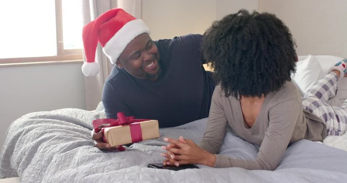 African American couple exchanging gift, woman unwrapping box and embracing on bed under lights
