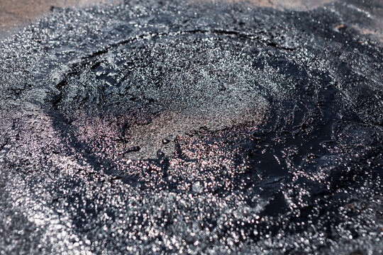 Roof repair in progress, freshly laid circular patch of glossy black asphalt sealant applied over damaged shingles to prevent leaks. Textured roofing material in darker grays with subtle overlaps 