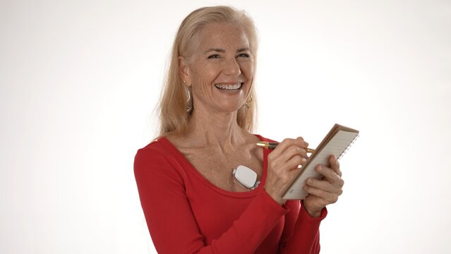 Mature woman wears a heart monitor, smiling while writing notes in a notebook, emphasizing health management and wellness