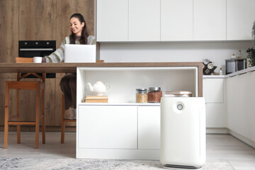 Young woman using laptop with cup of tea at table and air purifier in kitchen