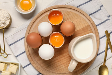 Cutting board with fresh chicken eggs, jug of milk and flour on light wooden table