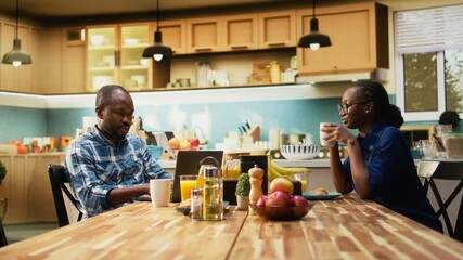 African American couple working from home together in the kitchen during the morning. Man types on laptop and woman enjoys coffee, symbolizing remote work and a balanced lifestyle. Camera A.