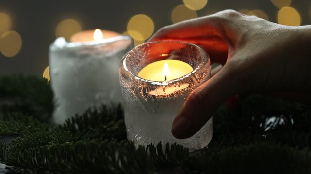 DIY Christmas lantern. Woman taking burning ice candle and fir branches at table against grey background with blurred lights, closeup. Bokeh effect