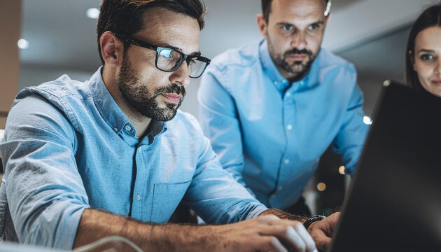 Focused Team Collaboration: In the hushed glow of late-night office work, focused colleagues intently review project details on a laptop, exhibiting dedication and partnership.