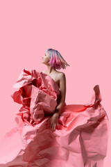 Model gracefully interacts with large crumpled sheets of pink paper, creating an artistic and dynamic scene against a soft pink background. The movement captures elegance and playfulness