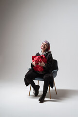 A person with purple hair smiles brightly while seated in a stylish chair. They hold a large red gift close, expressing excitement and joy in a simple, minimalist setting