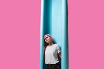 A young woman with pastel hair playfully leans against bold blue panels, surrounded by a striking pink backdrop. Her thoughtful expression invites creativity and whimsy