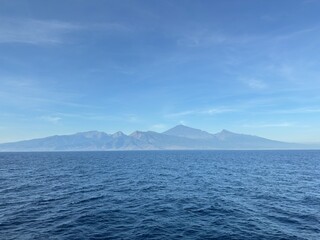 Vast blue ocean meets distant majestic mountain range under clear sky, evoking serenity and adventure for travel and nature lovers