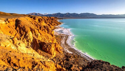 A stunning landscape featuring a bright turquoise lake with white salt deposits along its shore, bordered by dramatic orange-hued cliffs and distant mountains.