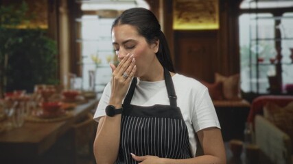 Young brunette woman wearing striped apron with hand on throat for choking in restaurant dining room; concern.